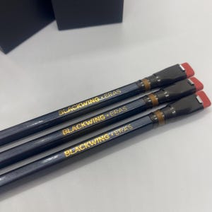 Peut inclure: Trois crayons gris foncé avec l'inscription dorée « BLACKWING ERAS ». Les crayons ont une bande marron et une gomme rouge. Les crayons sont disposés en diagonale, les gommes étant orientées vers le haut à droite.