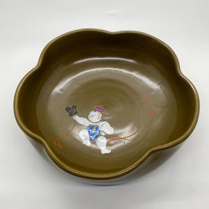 May include: A brown ceramic bowl with a scalloped edge. The interior features a painted illustration of a small figure in a blue diaper and a red and blue hat, holding a black object with red streamers. Three small red shapes are also painted inside the bowl.