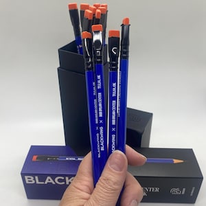 Può includere: Matite blu con gomme arancioni tenute in mano. Le matite sono stampate con "BLACKWING x BOB DYLAN CENTER TULSA, OK" e "LIFE IS ABOUT CREATING YOURSELF". Sullo sfondo c'è una scatola nera.