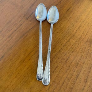 Puede incluir: Dos cucharas plateadas con mangos largos y cuencos ovalados descansan sobre una superficie de madera. Las cucharas tienen una superficie brillante y reflectante. Cada cuchara tiene un diseño decorativo en la base del mango. Las cucharas son probablemente para servir o comer.