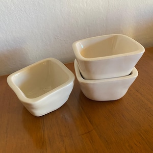 May include: Three small, off-white square ceramic bowls. Two bowls are stacked, and one is placed separately. The bowls have a slightly irregular shape and a glossy finish. They are arranged on a wooden surface.