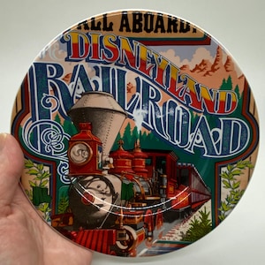 Puede incluir: Un plato redondo de cerámica con una imagen colorida de un tren de vapor vintage. El tren es rojo, verde y negro con detalles dorados. El texto "All Aboard Disneyland Railroad" está impreso en una fuente colorida y estilizada.