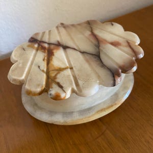 May include: A polished stone trinket box shaped like a seashell. The lid features a scalloped edge with carved grooves and a natural marbled pattern in shades of cream, beige, and deep brown. The base is a smooth, oval-shaped stone container.