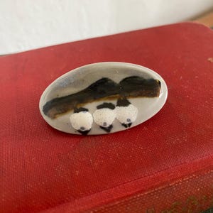 以下が含まれることがあります： 風景デザインの楕円形セラミックブローチ。ブローチは、濃い茶色の水平線と、黒いアクセントが付いた3つの白く斑点のある形状を持つ灰色の背景が特徴です。ブローチは赤い背景に配置されています。