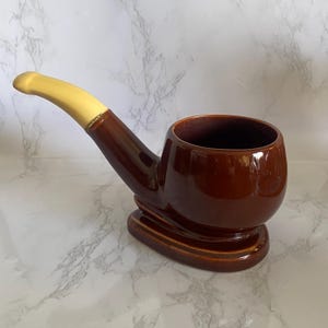 May include: A ceramic pipe-shaped mug featuring a glossy dark brown finish on the bowl and base, contrasted by a smooth, matte light yellow stem. The vessel has a wide, open circular top and sits on a flat, oval-shaped base for stability.