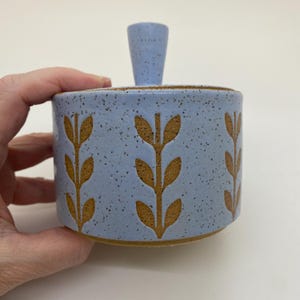 May include: A light blue ceramic jar with a lid and handle. The jar features a speckled finish and is decorated with brown leaf-like patterns. The jar is held by a hand against a white background. The jar is approximately 4 inches tall.