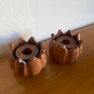 May include: Two wooden candle holders, each carved in a flower shape. The holders are a warm brown colour and have a circular indentation in the centre to hold a candle. The candle holders are on a wooden surface.