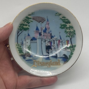 May include: A small white ceramic dish with a gold rim. The dish features a colorful illustration of Sleeping Beauty's castle at Disneyland, with the words "Disneyland" in gold lettering.