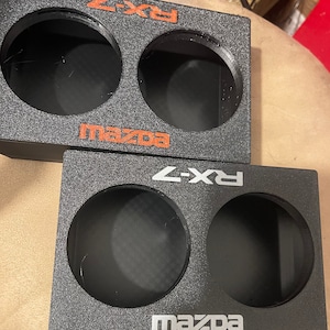 Puede incluir: Dos cajas de altavoces negras con dos aberturas circulares cada una. El texto "RX-7" y "MAZDA" está impreso en naranja y plata. Las cajas tienen una superficie texturizada.