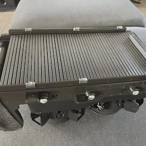 Puede incluir: Una parrilla rectangular negra con barras paralelas elevadas. La parrilla está hecha de un material oscuro y tiene varios pernos y sujetadores de color plateado. La parrilla está sobre una superficie gris.