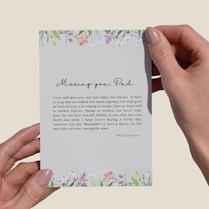 Op de afbeelding: Een rechthoekige kaart met de tekst "Missing you, Dad" en een oprechte boodschap. De kaart is omrand met aquarel bloemenillustraties in roze, paarse en gele tinten. De kaart wordt door twee handen vastgehouden.