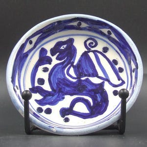 May include: A small, round ceramic dish with a hand-painted blue dragon design on a white background. The rim features a blue and white pattern. The dish is displayed on a black metal stand.