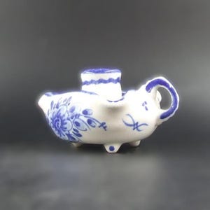 May include: A white ceramic creamer with blue floral patterns and a blue handle. The creamer has a small spout and stands on three small feet. A small, cylindrical container is attached to the top.
