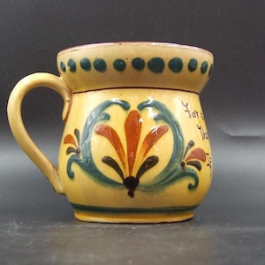 May include: A yellow ceramic mug with a handle, decorated with a floral design in shades of brown, green, and blue. The rim features a series of green dots. The mug has text on the side.