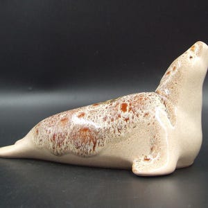May include: A ceramic seal ornament in a reclining position. The seal has a light beige body with brown speckled markings. The head is raised, and the ornament is set against a dark background.