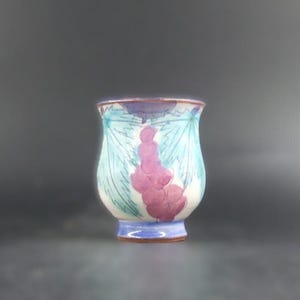 May include: A hand-painted ceramic cup with a flared rim. The cup features a white base with a design of purple grapes and blue leaves. The rim and base are trimmed with blue and purple paint. The cup is approximately 10 cm tall.