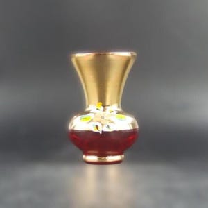 May include: A small decorative vase with a gold rim and base. The body of the vase is a deep red colour, with a gold floral design around the middle. The vase is approximately 13 cm tall.