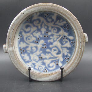 May include: A round ceramic dish with a light blue interior featuring a white floral pattern and small brown dots. The rim is a speckled beige colour, and it has two small handles on either side.