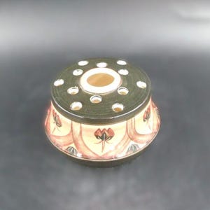 May include: A ceramic flower frog with a dark green top with multiple holes for arranging stems. The sides are decorated with a repeating pattern of red and brown floral motifs against a cream background. The base is dark green.