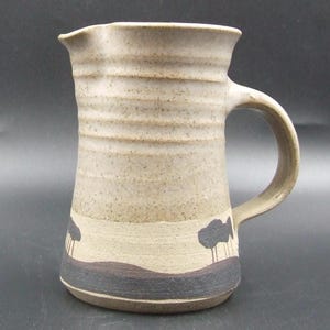 May include: A handmade ceramic jug with a beige speckled finish and a curved handle. The jug features a landscape design with black trees and a dark brown base. The top has a slightly irregular rim.