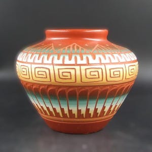 May include: A terracotta-coloured ceramic vase with a rounded body and a small opening. The vase features geometric patterns in yellow, turquoise, and white. The design includes repeating square spirals and stepped lines, with a textured, carved appearance.