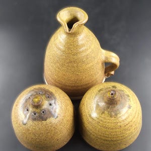 May include: A set of three handcrafted ceramic items in shades of brown and tan. The set includes a jug with a handle and two round salt or pepper pots with multiple holes on top. The pottery has a rustic, textured appearance.