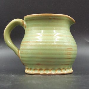 May include: A light green ceramic jug with a curved handle and a spout. The jug features horizontal ridges and a decorative scalloped base. The rim and base have a brown trim. It's a rustic, handcrafted piece, perfect for serving.