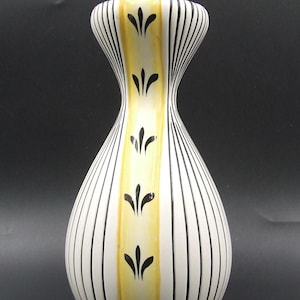 May include: A white ceramic vase with black vertical stripes and a central yellow panel. The yellow panel is decorated with black leaf-like motifs. The vase has a slender neck and a bulbous base, creating an elegant silhouette.