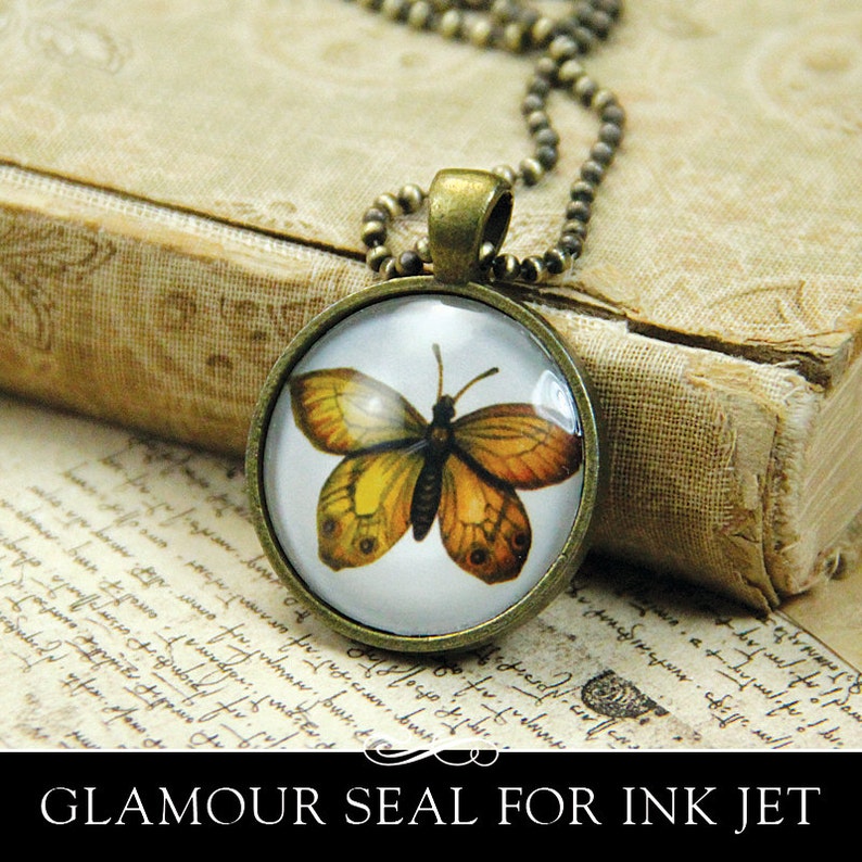 May include: A gold chain necklace with a round glass pendant featuring a vintage illustration of a butterfly with brown and yellow wings. The pendant is set against a white background. The necklace is resting on a vintage book with handwritten text.