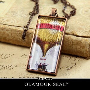 May include: A copper-toned pendant necklace with a rectangular glass charm featuring a vintage illustration of a hot air balloon with the word "ACHA MB" printed on a red banner.