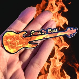 May include: A fiery orange and yellow bass guitar sticker with the text "Bass Is Boss" in black. The sticker is held in a hand against a background of flames.