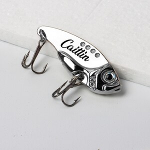 May include: A silver-colored fishing lure in the shape of a fish, with two treble hooks and the name "Caitlin" printed on the side. The lure has a reflective surface and a detailed eye.