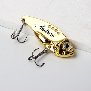 May include: A gold-colored fishing lure with the name "Andrew" in black script. The lure has a fish-like shape with a detailed eye and several small holes. It features two treble hooks.