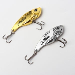 May include: Two personalized fishing lures. One is gold-colored with the name "Andrew" in black script. The other is silver-colored with the name "Caitlin" in black script. Both lures have a fish-shaped design and include two treble hooks.