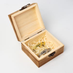 May include: A wooden box with a hinged lid, open to reveal two fishing lures nestled in shredded paper. One lure is gold with the name "Andrew", the other is silver with "Caitlin". The box is made of light-colored wood.