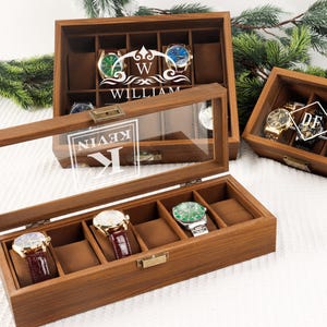 Peut inclure: Coffrets en bois pour montres avec couvercles en verre. Les coffrets sont bruns et contiennent diverses montres avec des cadrans et des bracelets de différentes couleurs. Certains coffrets ont du texte personnalisé, notamment des noms et des initiales.