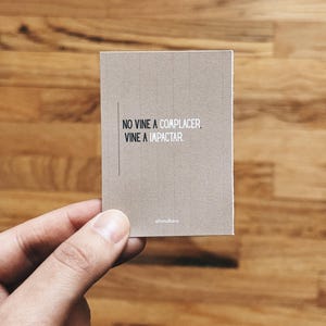 May include: A small, rectangular card with the text "NO VINE A COMPLACER, VINE A IMPACTAR." in black font. The card is a light brown color and is held by a hand. The brand name "aliandkaro" is printed at the bottom.