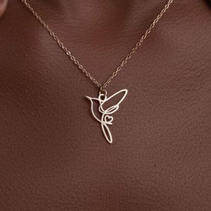 Puede incluir: Un delicado collar dorado con un colgante de colibrí. El colgante es un diseño minimalista de línea continua con un detalle de corazón. El collar se muestra en el pecho de una persona, con las manos visibles a ambos lados.