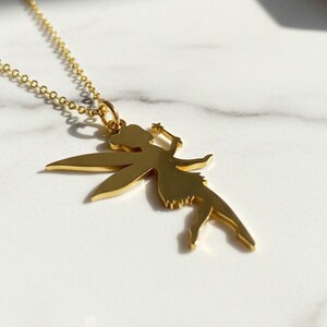 May include: A gold-coloured necklace with a fairy pendant. The fairy has outstretched wings and holds a wand. The chain is delicate, and the pendant is detailed. The necklace is displayed on a white marble surface.