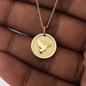 May include: A gold-colored necklace featuring a circular pendant with a dove carrying an olive branch. The pendant is suspended from a delicate chain. The dove is in flight, and the overall design is simple and elegant.
