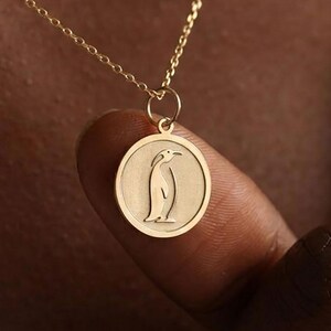 May include: A gold necklace with a circular pendant showing a penguin design. The pendant hangs from a fine chain. The penguin is illustrated in a simple, elegant style, making it a charming piece of jewellery.