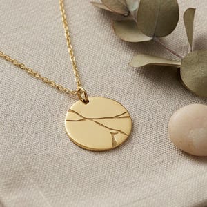 Op de afbeelding: Een goudkleurige ketting met een ronde hanger met een gebarsten ontwerp. De ketting ligt op een beige linnen doek, vergezeld van een kleine steen en een takje groen. De ketting is delicaat en de hanger is glad.