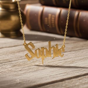 May include: A gold-colored necklace with the name "Sophie" in a stylized font. The necklace hangs in front of a wooden surface, with a quill, inkwell, and leather-bound books in the background. The name is the focal point of the image.