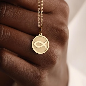 Könnte beinhalten: Eine goldfarbene Halskette mit Anhänger, die einen kreisförmigen Anhänger mit einem gravierten Ichthys-Symbol, auch bekannt als Jesusfisch, aufweist. Der Anhänger hängt an einer filigranen Kette. Die Halskette wird vor dunkler Haut gehalten.