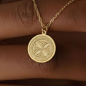 Puede incluir: Un collar dorado con un colgante circular. El colgante presenta un intrincado diseño de nudo celta dentro de un marco circular. La cadena del collar es delicada y fina, y el colgante está suspendido de ella.