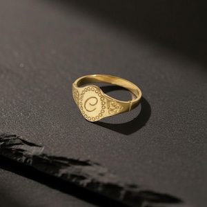 Peut inclure: Bague chevalière dorée avec la lettre "C" gravée sur le dessus. La bague présente un motif de bordure circulaire détaillé. Posée sur un fond sombre, elle est un bijou classique et élégant.