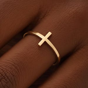 May include: A gold ring with a cross design. The ring has a simple band and a cross-shaped feature. The ring is made of a shiny, metallic material. The cross is positioned centrally on the ring.