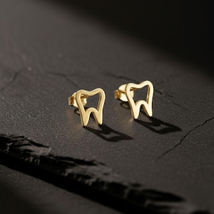 Könnte beinhalten: Ein Paar goldfarbene Ohrstecker in Form eines Zahns. Die Ohrringe sind vor einem dunklen Hintergrund platziert, wobei Schatten Tiefe verleihen. Das Design ist schlicht und modern, passend für Zahnärzte oder Interessierte der Zahnmedizin.