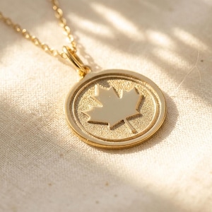 May include: A gold-colored necklace featuring a circular pendant with a detailed maple leaf design. The pendant is suspended from a delicate gold chain. The necklace is displayed on a textured, off-white surface.