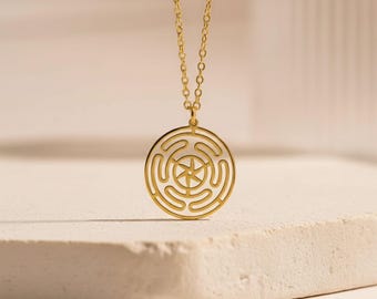 May include: Gold-colored pendant necklace featuring a circular design with a maze-like pattern and a starburst center. The pendant hangs from a delicate gold chain. The necklace is displayed on a neutral-colored surface, highlighting the jewelry's design.
